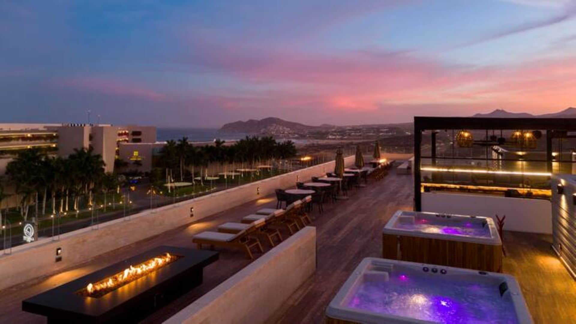Une terrasse sur le toit avec des sièges extérieurs, un foyer et un jacuzzi, offrant une vue sur le paysage urbain et les montagnes au crépuscule.