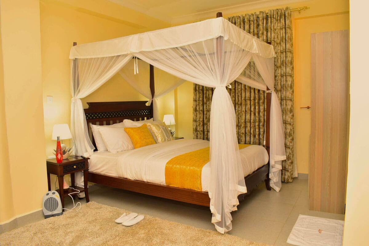 A four-poster canopy bed with white bedding and a yellow throw, a bedside table with a lamp, and a red decorative item.