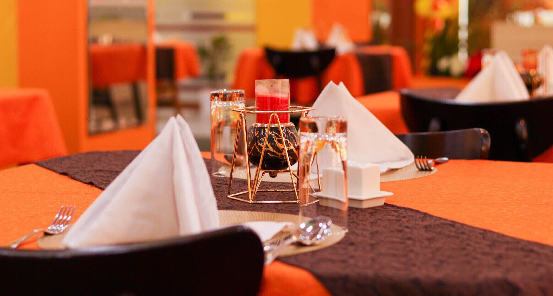 Eettafel gedekt met een servet, een glas en een klein bordje, in een restaurant met oranje stoelen en tafels.
