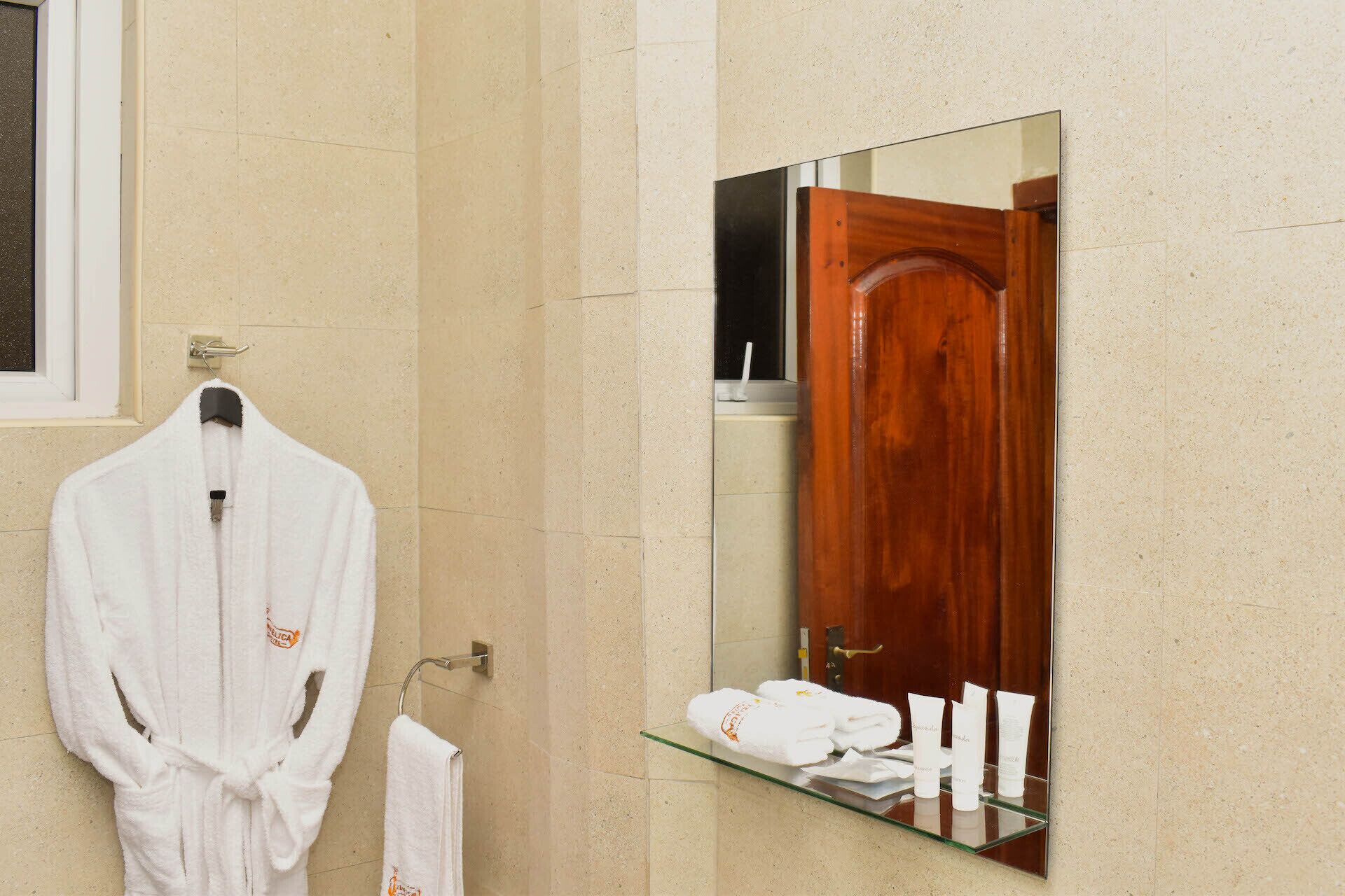 Une salle de bain avec un peignoir blanc suspendu à un porte-serviettes, une étagère en verre contenant des articles de toilette et une porte en bois qui se reflète dans un miroir.