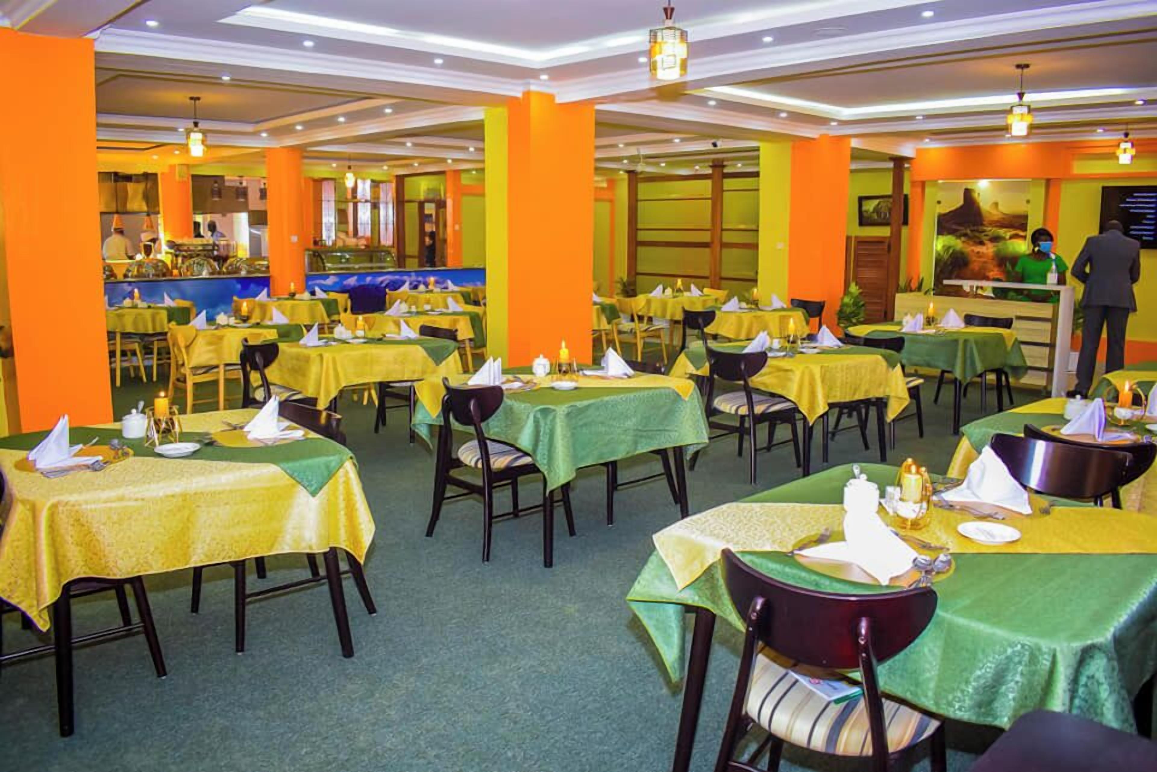 A restaurant with multiple tables covered in yellow tablecloths, green chairs, and orange pillars.