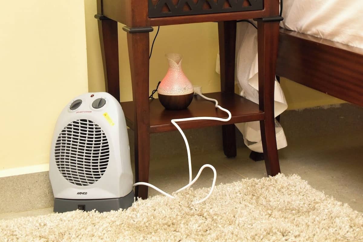 A bedroom with a wooden nightstand, a lamp, a red vase, a white lamp shade, a bed with a white canopy, and a portable heater.