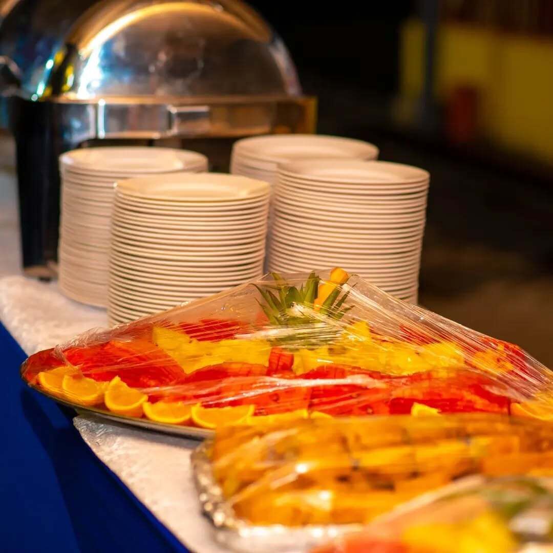 A buffet setup with stacked white plates and a covered dish with sliced fruits.