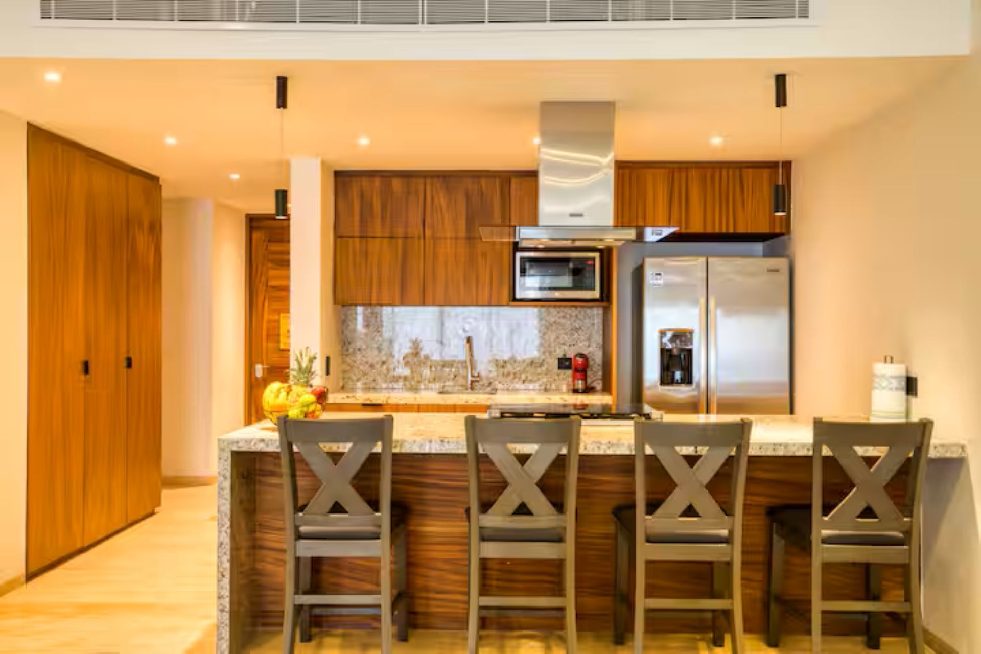Cocina moderna con armarios de madera, refrigerador de acero inoxidable, microondas y encimera de granito. Hay cuatro taburetes de bar con un diseño original en forma de X.