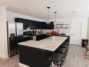 Una cocina moderna con gabinetes negros, electrodomésticos de acero inoxidable, una isla central con taburetes de bar, iluminación colgante y un salpicadero con diseño de espiga.