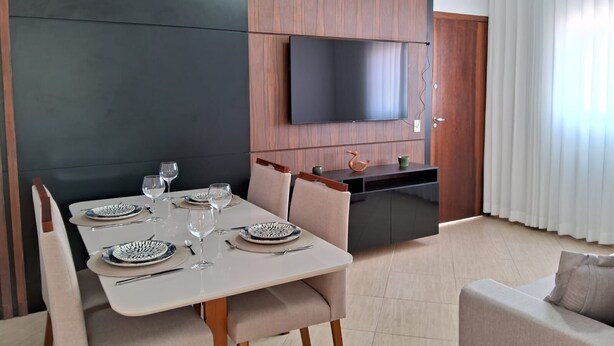 A modern living room with a dining area, a flat-screen TV mounted on the wall, and a kitchen area with a wooden cabinet. - Apto Prédio Costa 1 c/ garagem no centro (São Roque)