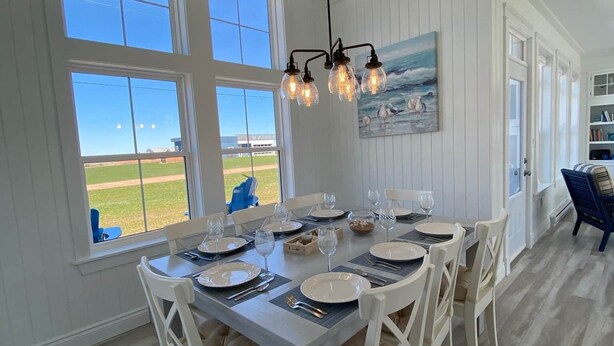 A dining area with a large table set for six, white chairs, and a painting on the wall. - Blue Horizon Cottage   – Steps From Private Beach, Family-Friendly Coastal Retreat (Prince Edward Island)