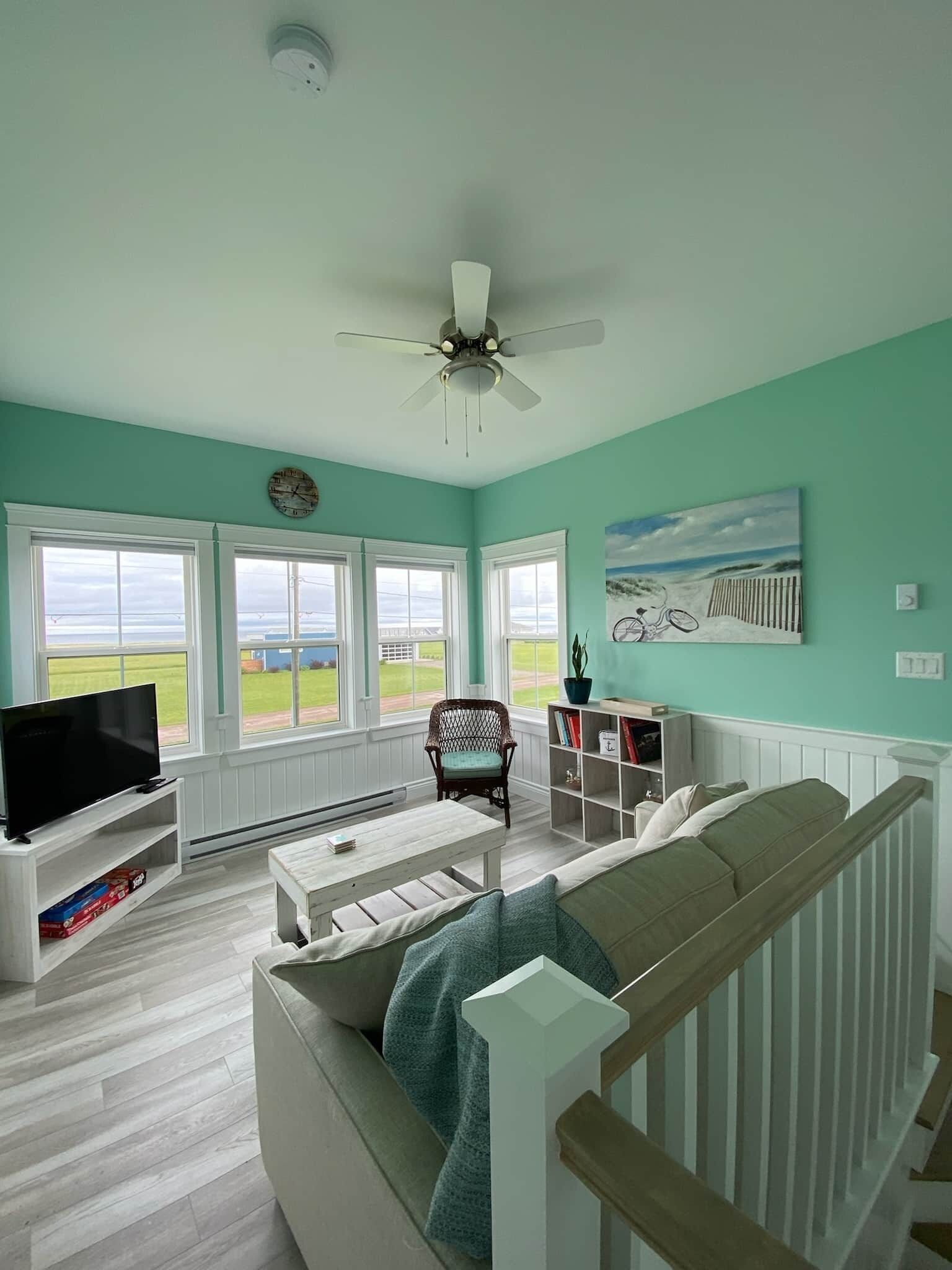 A living room with a large window, a ceiling fan, a television, a bookshelf, and a painting.