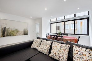 Un salon moderne avec un canapé noir, des coussins à motifs floraux, un coin repas avec une nappe rouge et une grande fenêtre donnant sur des immeubles.