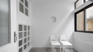 A hallway with two white chairs, a door with a metal handle, and a window with a view of the outside.