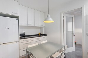 Moderni keittiö, jossa on valkoiset kaapit, musta työtaso, ruostumattomasta teräksestä valmistettu jääkaappi ja lasipöytä.