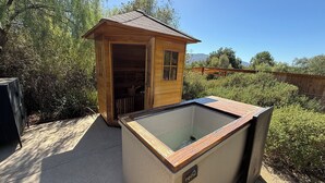 A wooden cabin with a glass-enclosed hot tub, surrounded by greenery and a clear sky.