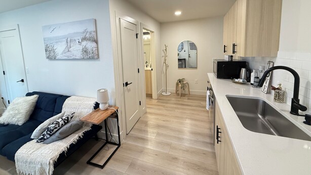 A modern apartment with a living area featuring a sofa, a coffee table, and a painting. The kitchen has a white countertop, a sink, and wooden cabinets. - The Sable Stay (Vancouver)