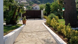 Un chemin pavé mène à une entrée fermée par un portail, d’où l’on peut admirer une colline et des immeubles résidentiels.