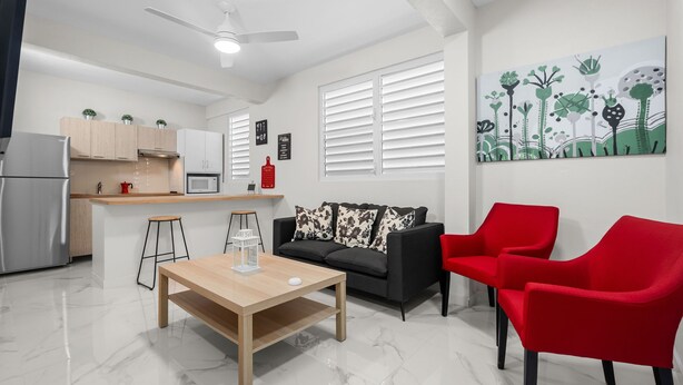 A modern living space with a kitchen, a dining area, and a seating area with a sofa and red chairs. - Modern 2 BR Apt Great Location Smart TV AC (Manati)