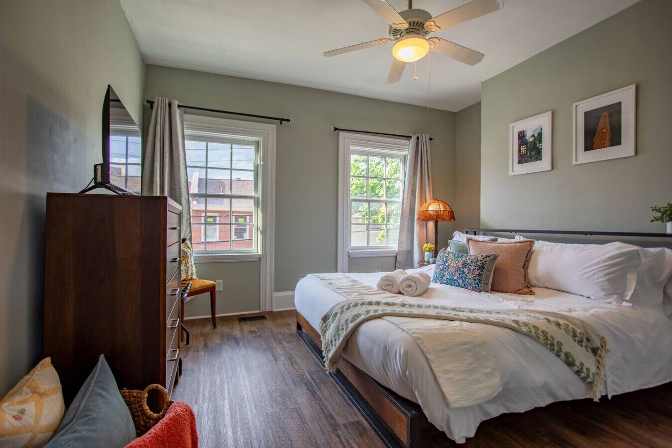 Une chambre à coucher comprenant un lit, une commode, une chaise, un ventilateur de plafond et deux fenêtres.