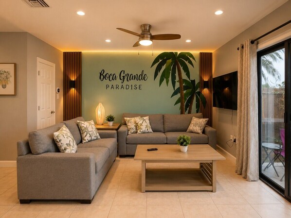 Un salon comprenant un canapé, une table basse, un ventilateur de plafond, un téléviseur fixé au mur et une grande plante.