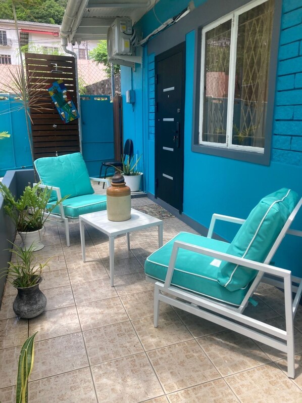 A blue-painted outdoor patio with white furniture, including two chairs and a table, surrounded by potted plants.