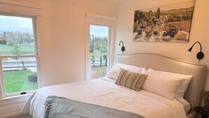 Une chambre à coucher avec un grand lit, deux fenêtres, un tableau représentant des bouteilles de vin et une table de chevet avec une lampe.