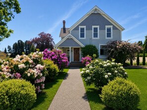 Een tweelaags huis met een veranda, een pad naar de voordeur en een goed onderhouden tuin met verschillende bloeiende planten.