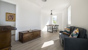 Een moderne woonkamer met een bank, een houten kast, een eettafel met stoelen en een groot raam dat voor natuurlijk licht zorgt.