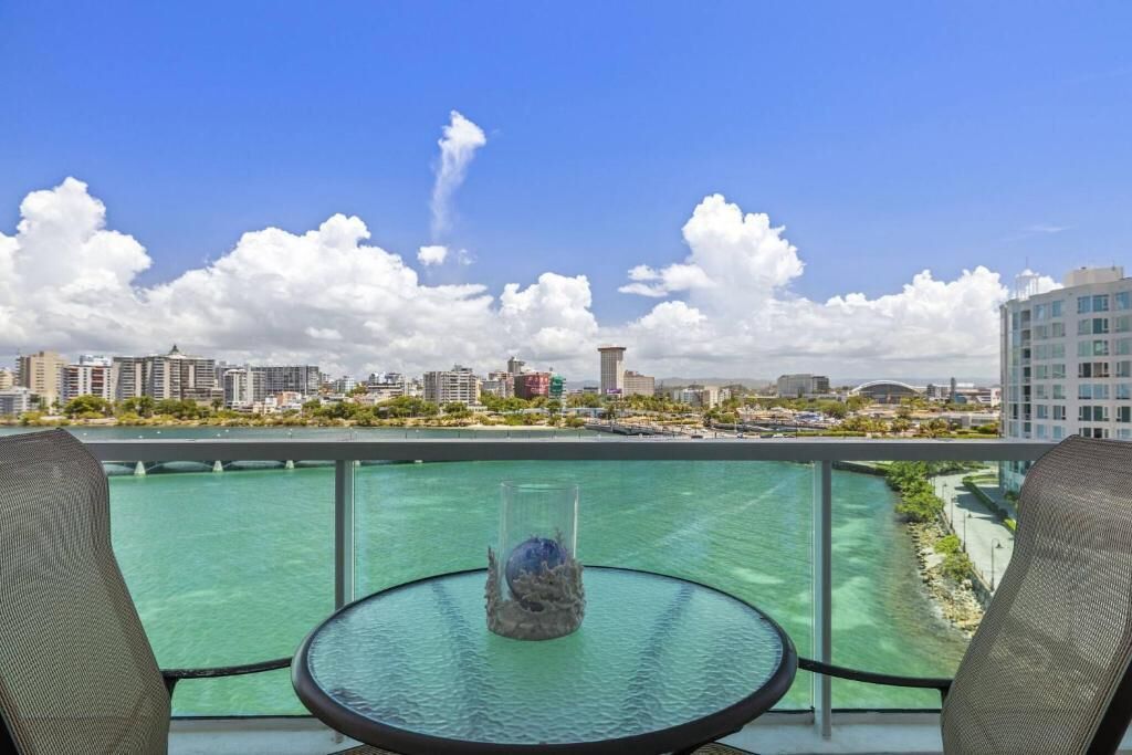 A balcony with a glass table and two chairs, overlooking a body of water and a cityscape with buildings and a bridge.