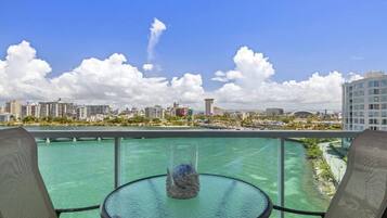 A balcony with a glass table and two chairs, overlooking a body of water and a cityscape with buildings and a bridge.