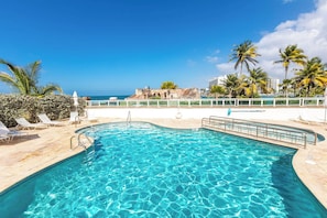 A clear blue pool with lounge chairs, palm trees, and a view of the ocean and buildings.
