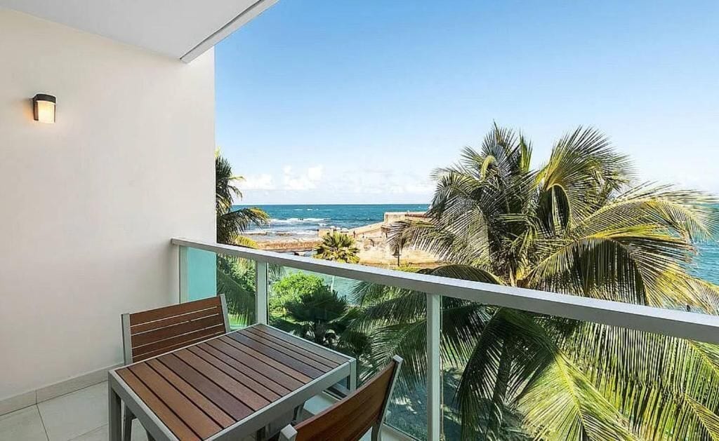A balcony with a wooden table and chairs, overlooking a beach with palm trees and the ocean.