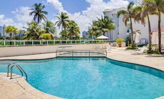 Una piscina con una escalera de metal, rodeada de palmeras y un edificio blanco al fondo.