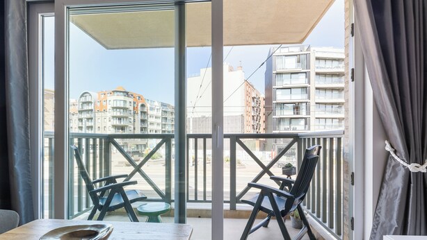 Ein Balkon mit Tisch und Stühlen, eine Schiebetür aus Glas und Blick auf Gebäude im Hintergrund.