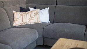 Ein graues Ecksofa mit drei Kissen, davon eines mit Palmenmuster, und ein Holztisch.