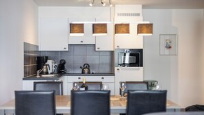 Eine moderne Küche mit weißen Schränken, einer Kaffeemaschine, einem Wasserkocher und einer Mikrowelle. Im Vordergrund steht ein Esstisch mit Stühlen.