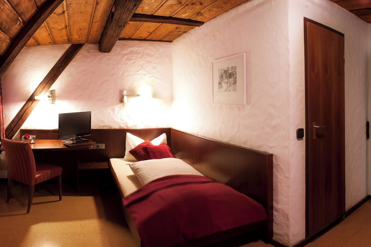 A bedroom with a bed, a desk with a computer, a chair, a wooden door, and a framed picture on the wall.