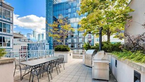 Eine Terrasse im Freien mit Tisch, Stühlen, einem Grill und Blick auf eine Stadtlandschaft mit modernen Gebäuden.