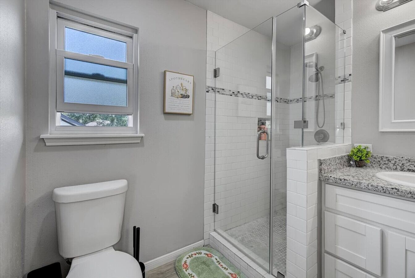A bathroom with a toilet, a shower with a glass door, a sink with a granite countertop, and a window with a view of the sky.