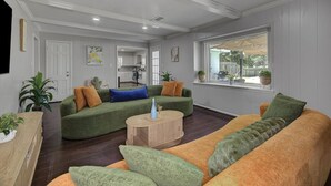 Un salon spacieux avec un canapé vert et orange, une table basse en bois et une cuisine en arrière-plan.