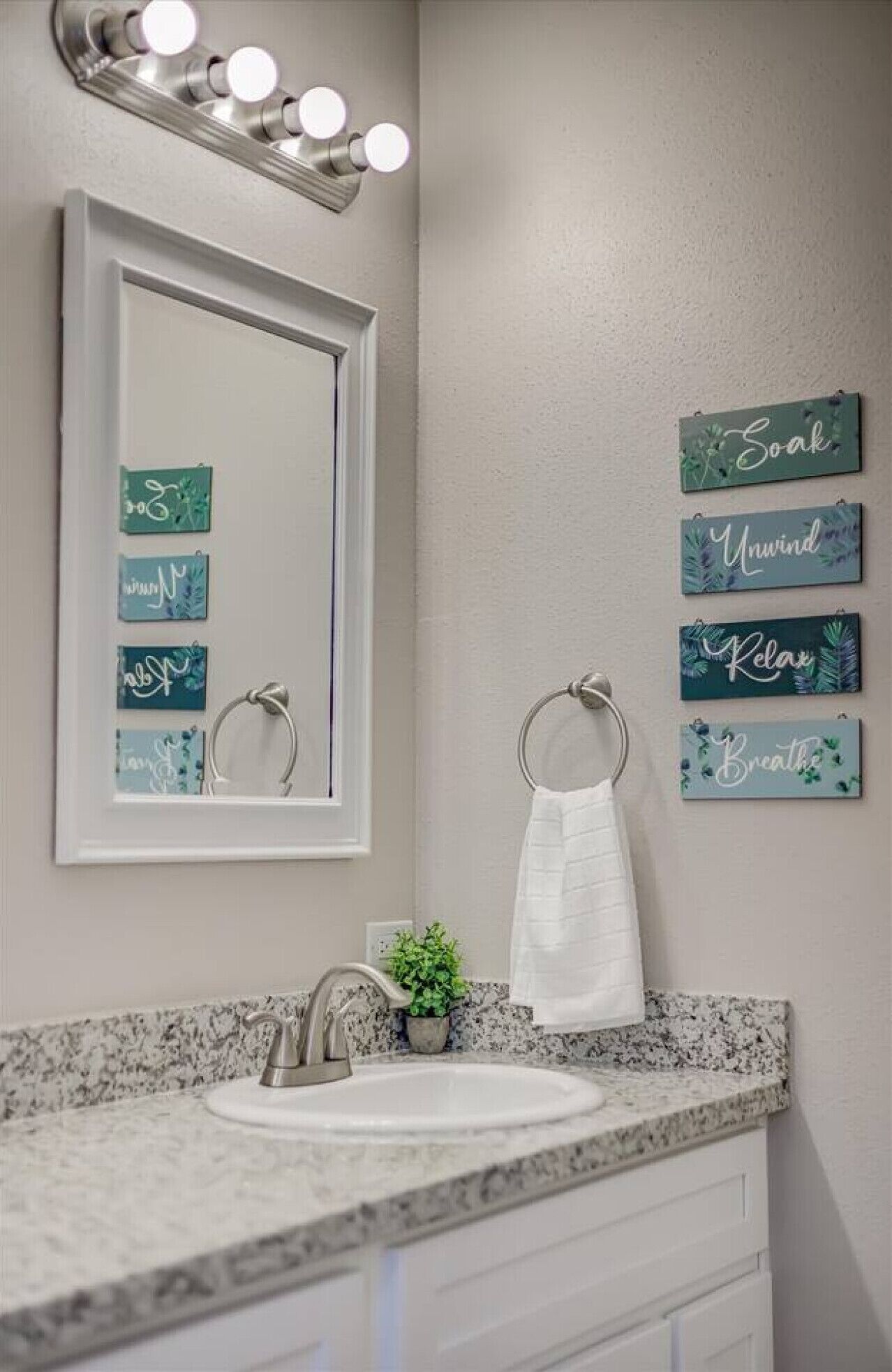 Une salle de bain avec un meuble-lavabo, un évier et un miroir. Des appliques murales sont fixées au-dessus du miroir, et quatre panneaux encadrés affichent des phrases motivantes.
