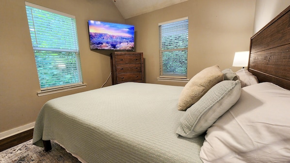 A bedroom with a bed, a dresser, a television on the wall, and two windows with blinds.