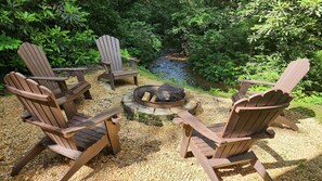 A stone fire pit with Adirondack chairs sits beside a running stream.