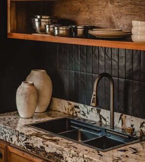 A kitchen with a granite countertop, a double sink, two ceramic vases, and a wooden shelf above holding bowls and plates.