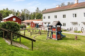 A playground with swings and a slide, surrounded by a grassy area and a building with a red roof. - Relax in peaceful and sunny Marmaverken apartment (Marmaverken)