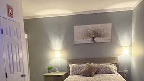 A bedroom with a bed, bedside lamps, a nightstand, a wall-mounted painting, and a door with a safety sign. - Palmas View 2 (catano)