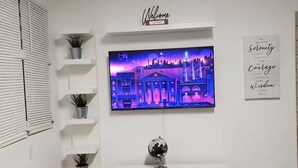A living room with a flat-screen TV mounted on the wall, a white entertainment unit below, and a small globe on the unit. There are potted plants on shelves to the left of the TV. - Palmas View 2 (catano)