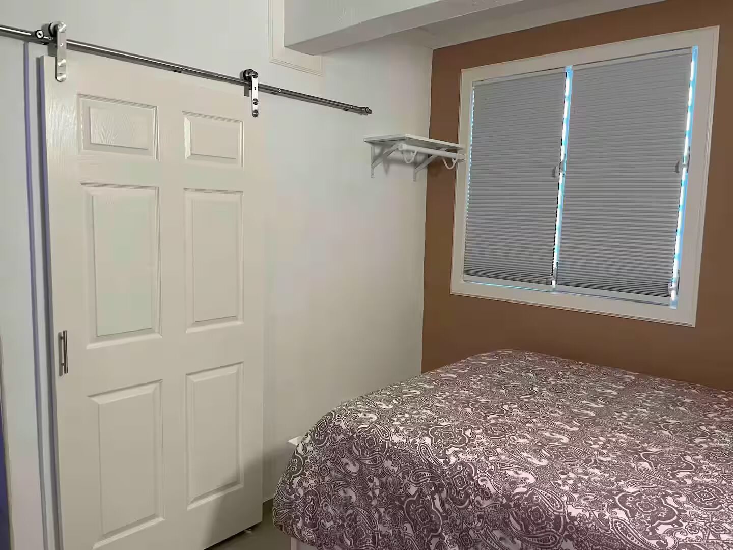 Un dormitorio con cama, una puerta, una ventana con persianas y una barra para cortinas en el techo.