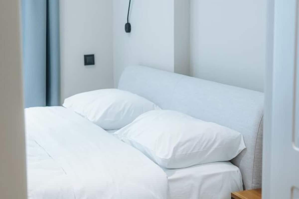 Un lit avec une literie blanche, dans une pièce dont un mur est peint en blanc et où se trouve une petite table de chevet.