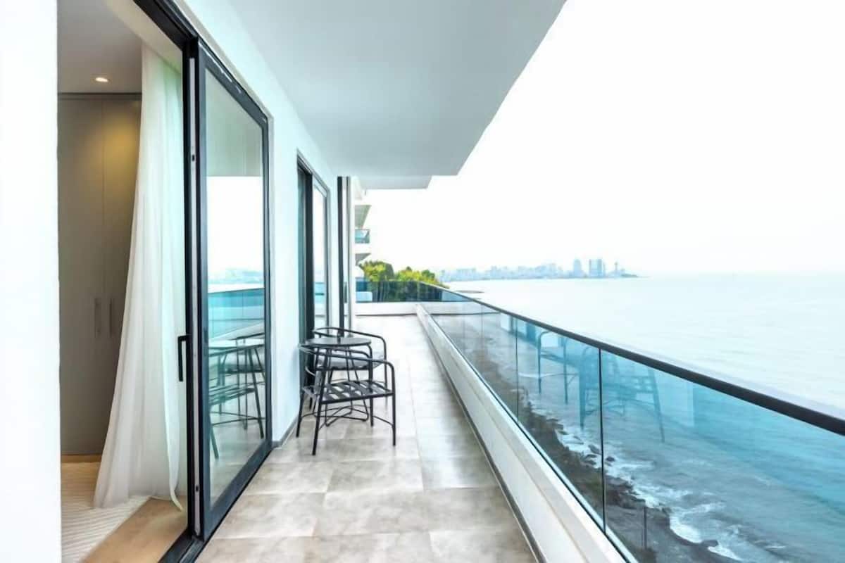 Un balcon doté d’une balustrade en verre donnant sur la mer, une table avec des chaises, et une vue sur les toits de la ville au loin.
