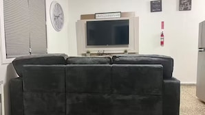 A living room with a large dark-colored sofa, a flat-screen TV mounted on the wall, and a small potted plant on the windowsill.