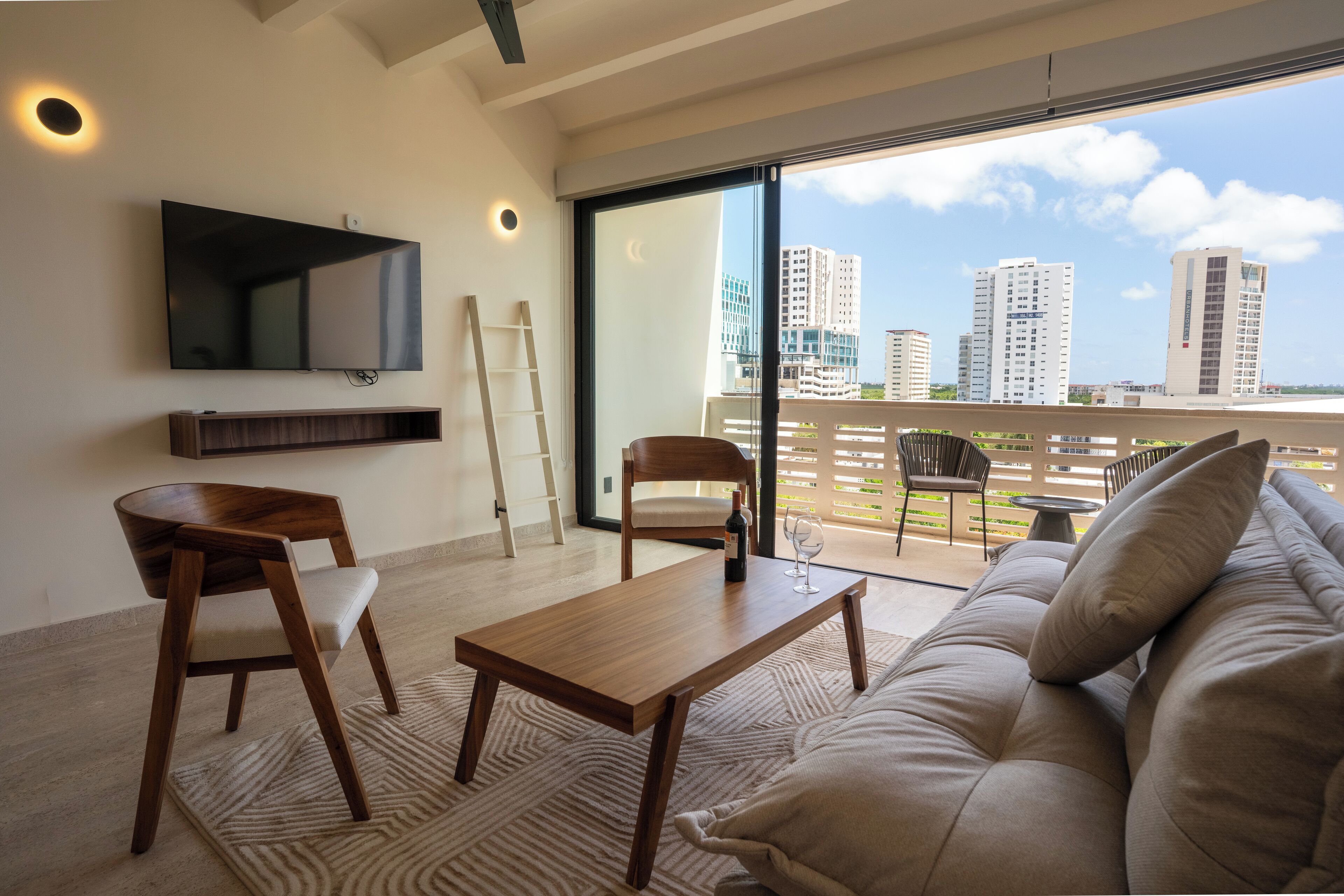 Un salon moderne avec un canapé, deux fauteuils en bois, une table basse et un téléviseur fixé au mur.