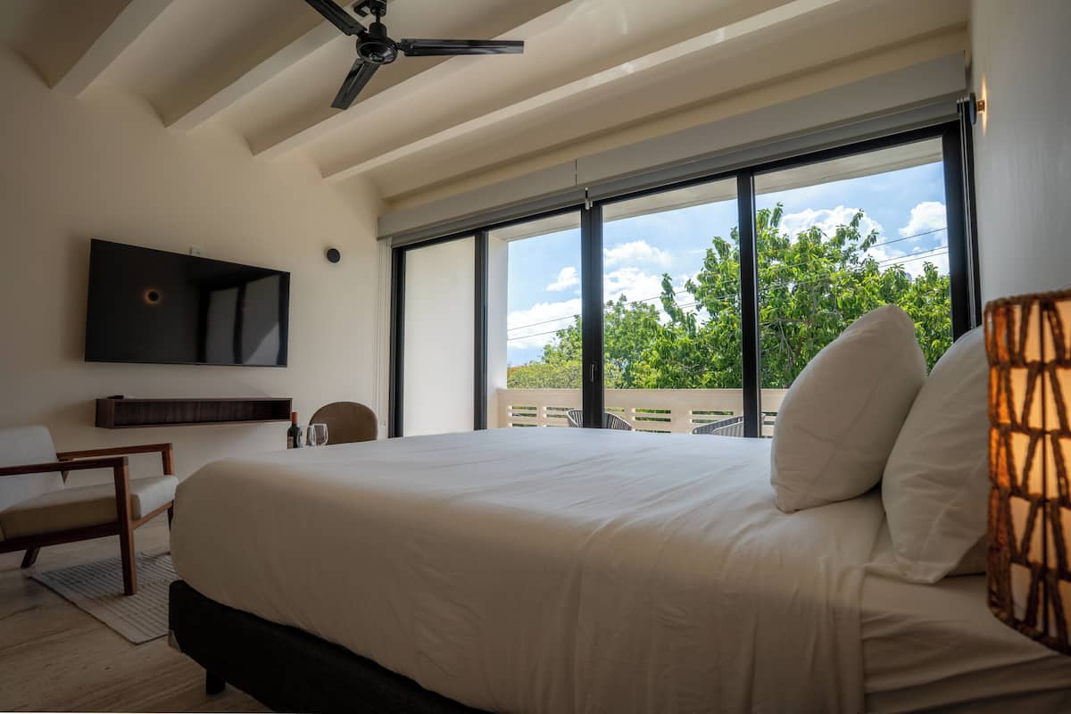 Quarto com uma cama grande, um ventilador de teto, uma televisão na parede e vista para o verde lá fora.